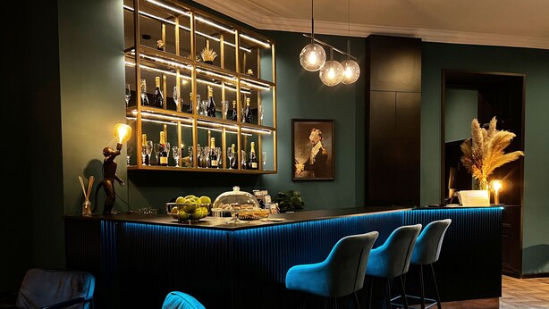 Un espace bar avec un comptoir bleu, de nombreux tabourets et une étagère où sont exposées des bouteilles et des verres.