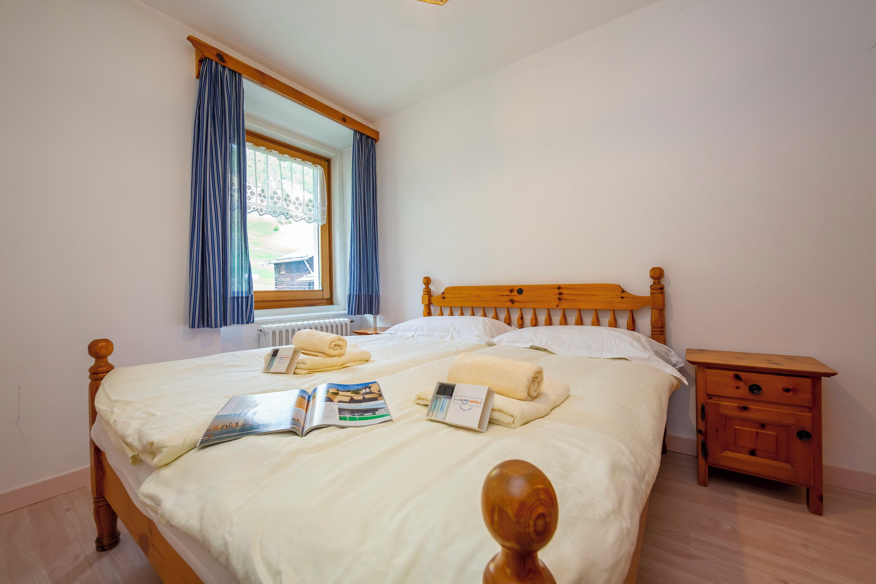 Un lit bien fait avec une tête de lit en bois, une petite table de chevet en bois dotée de tiroirs, une fenêtre avec des rideaux et un radiateur.