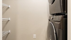 A laundry room with a stacked washer and dryer, a white wall, and a wooden floor. - Newly built and tastefully furnished legal one bedroom private guest suit. (Calgary)