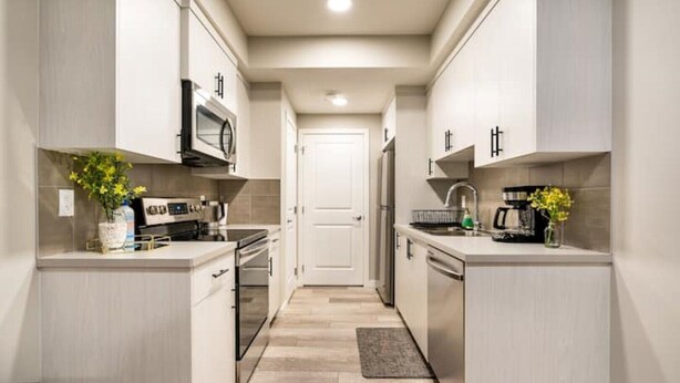 A narrow kitchen with white cabinets, a microwave, and a refrigerator. There are two sinks and a door at the end of the kitchen. - Newly built and tastefully furnished legal one bedroom private guest suit. (Calgary)