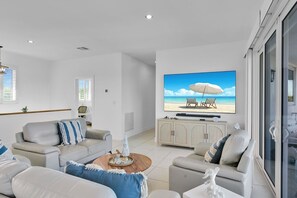 A modern living room with a large flat-screen TV, a sofa set, a coffee table, and a view of a beach through the window.