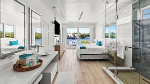 A modern hotel room with a large window, a shower, a bed, and a vanity with a sink.