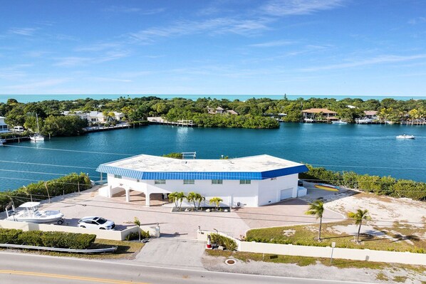 A white building with a blue roof, a parking area, and a view of a body of water with boats. - Paradise Cove on the Gulf 5 bedroom 3 bath with pool Ice Machince dockage, kayaks, bikes, paddleboards and more (Marathon)