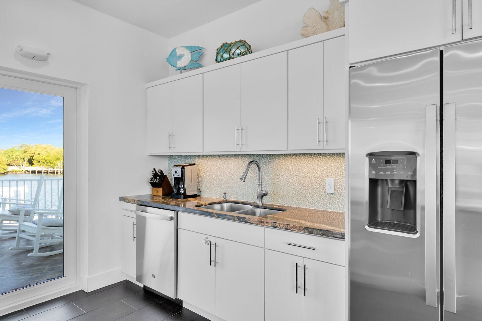 Una cocina moderna con armarios blancos, un refrigerador de acero inoxidable, un dispensador de agua integrado y vistas a un balcón con mobiliario exterior.