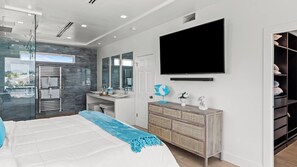 A modern bedroom with a large bed, a TV mounted on the wall, a bathroom with a glass shower enclosure, and a view of the outdoors.