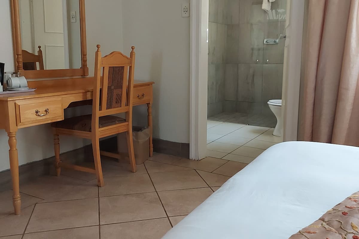 Quarto de hotel com escrivaninha de madeira, espelho grande, cama com colcha florida e área de chuveiro visível por uma porta de vidro.