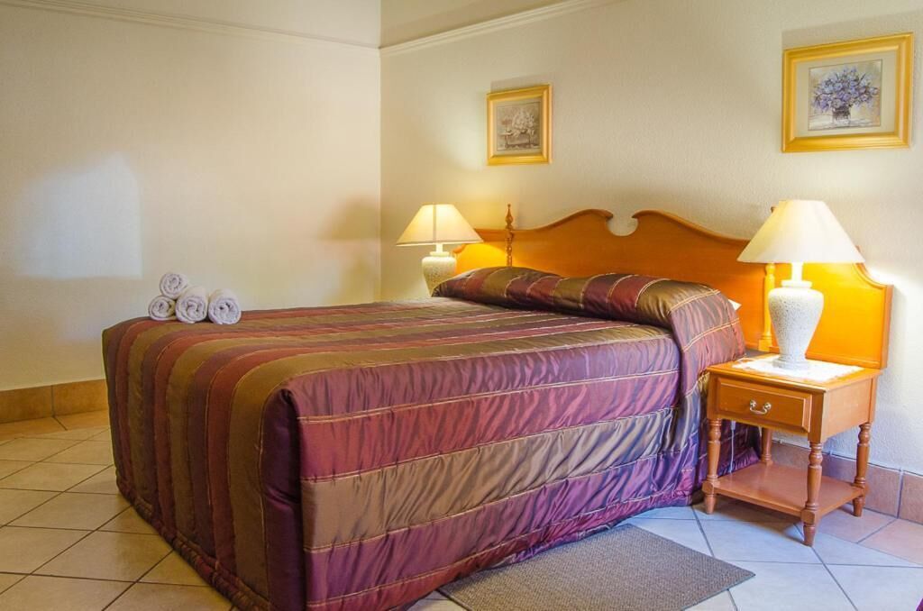 A hotel room with a bed, two bedside lamps, a wooden nightstand, and two framed pictures on the wall.