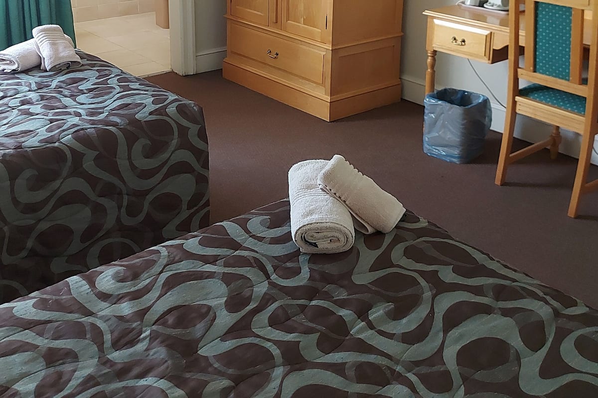 Quarto de hotel com duas camas, um guarda-roupa de madeira, uma escrivaninha com cadeira, um espelho e banheiro com chuveiro.