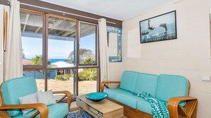 Une pièce avec un canapé de couleur turquoise, une chaise en bois et une table à manger sur laquelle se trouve un bol bleu. Une grande fenêtre offre une vue sur l’océan et sur un bâtiment dont le toit est rouge.