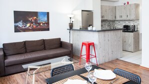 A modern living room with a brown sofa, a glass coffee table, and a dining area with a black table and chairs. A red stool is visible near the kitchen area, which includes wooden cabinets and a microwave.