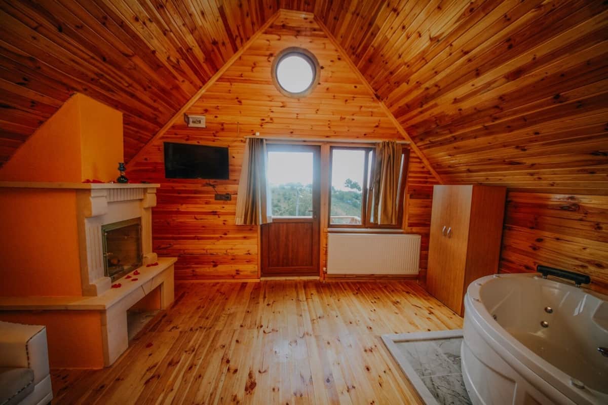 A cozy attic bedroom with a wooden sloped ceiling, a fireplace, a large window, and a bathtub.