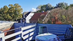 A rooftop terrace with a table covered in a patterned tablecloth, a blue bench, and a view of surrounding buildings and trees. - Vacation apartment (Moormerland)