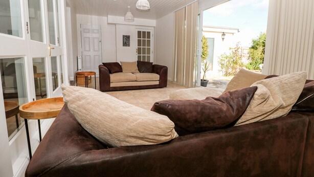 A spacious living area with a brown leather sofa, a beige sofa, and a wooden coffee table. There are two pendant lights hanging from the ceiling, and a sliding glass door leading to an outdoor area. - 5 Sea Way (Bognor Regis)