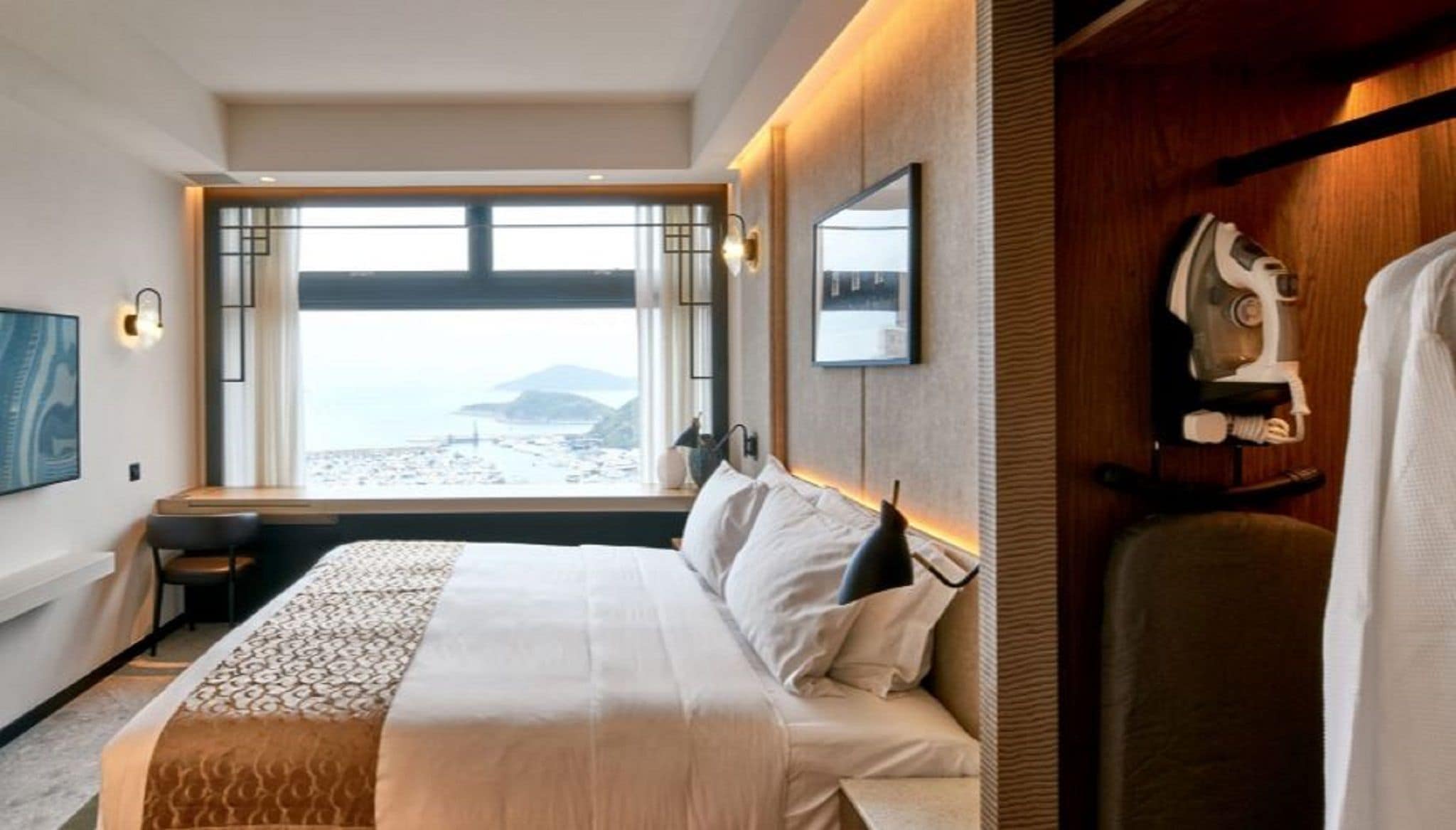 A hotel room with a large bed, a desk, a chair, a TV, and a view of the beach.