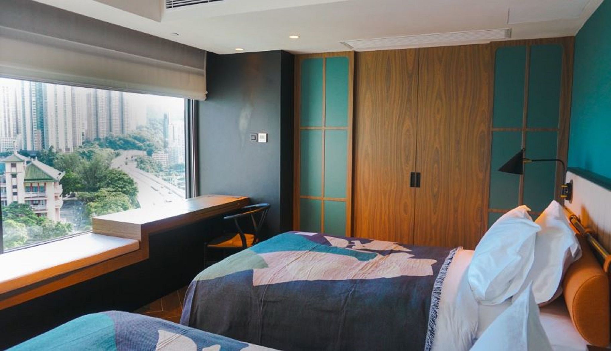A hotel room with a bed, a desk, a chair, a window with a view of buildings, and a wardrobe.