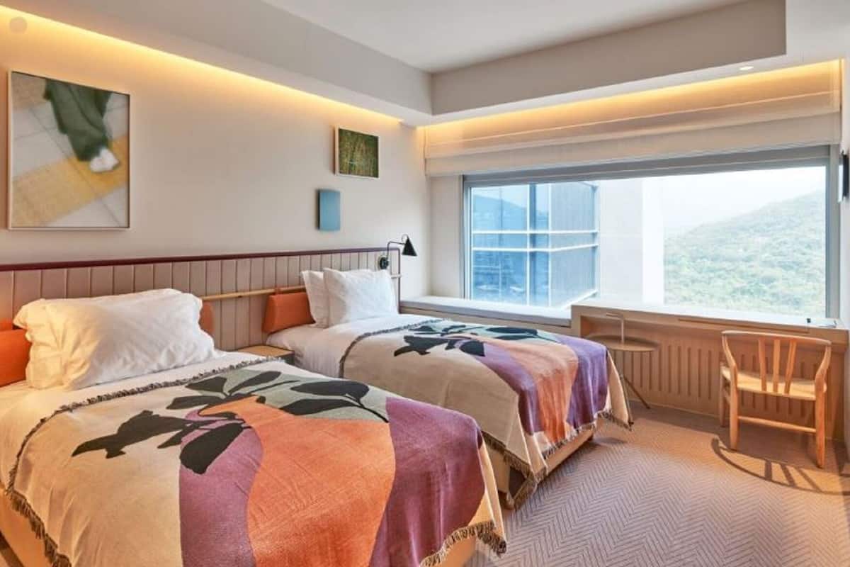 A hotel room with two beds, a desk, a chair, a window with a view, and artwork on the walls.