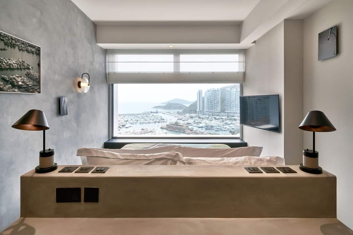 A modern bedroom with a large window, a bed, bedside lamps, and a flat-screen TV.