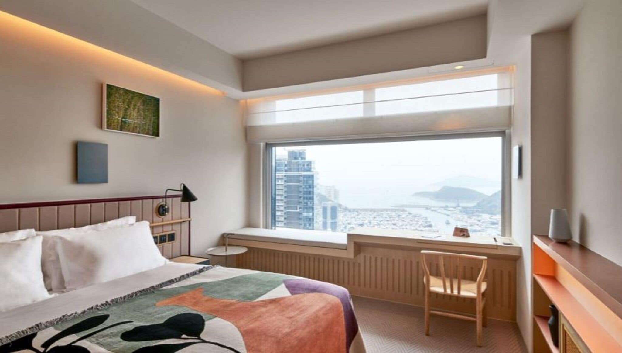 A hotel room with a bed, a desk, a chair, a window with a city view, and a radiator.