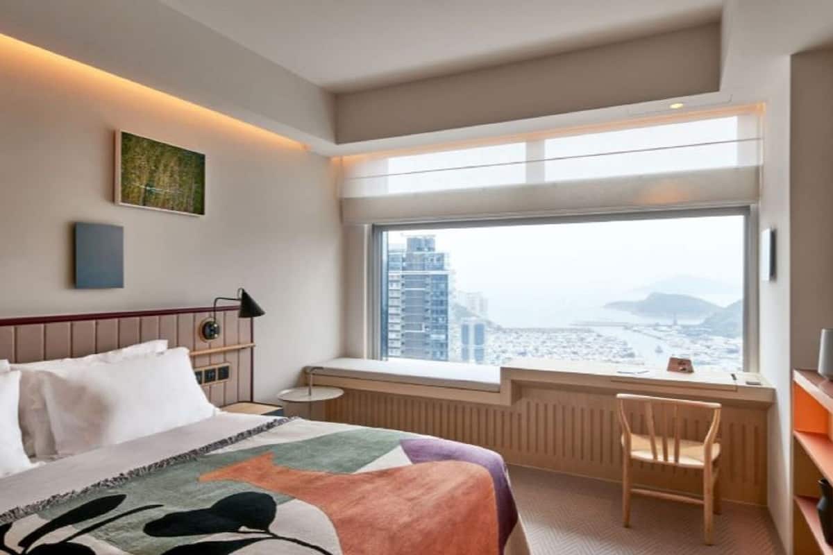 A hotel room with a bed, a desk, a chair, a window with a city view, and a radiator.