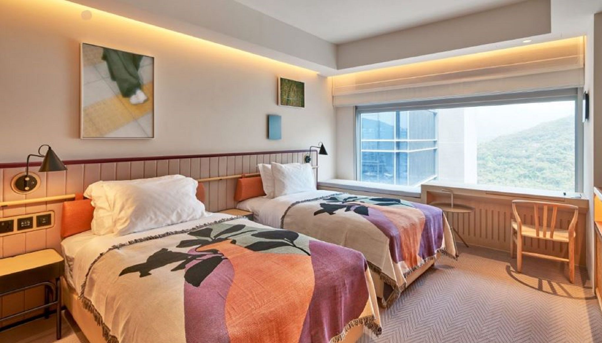 A hotel room with two beds, a desk, a chair, a window with a view, and artwork on the walls.