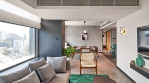 A modern living room with a sofa, dining table, and chairs, a large window with a city view, and a TV mounted on the wall.