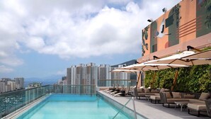 Piscina no terraço com espreguiçadeiras e guarda-sóis, cercada por edifícios altos e um céu limpo.