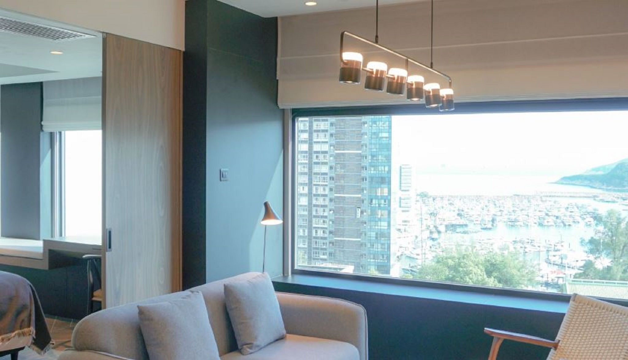 A modern hotel room with a large window, a sofa, a lamp, and a ceiling light fixture.