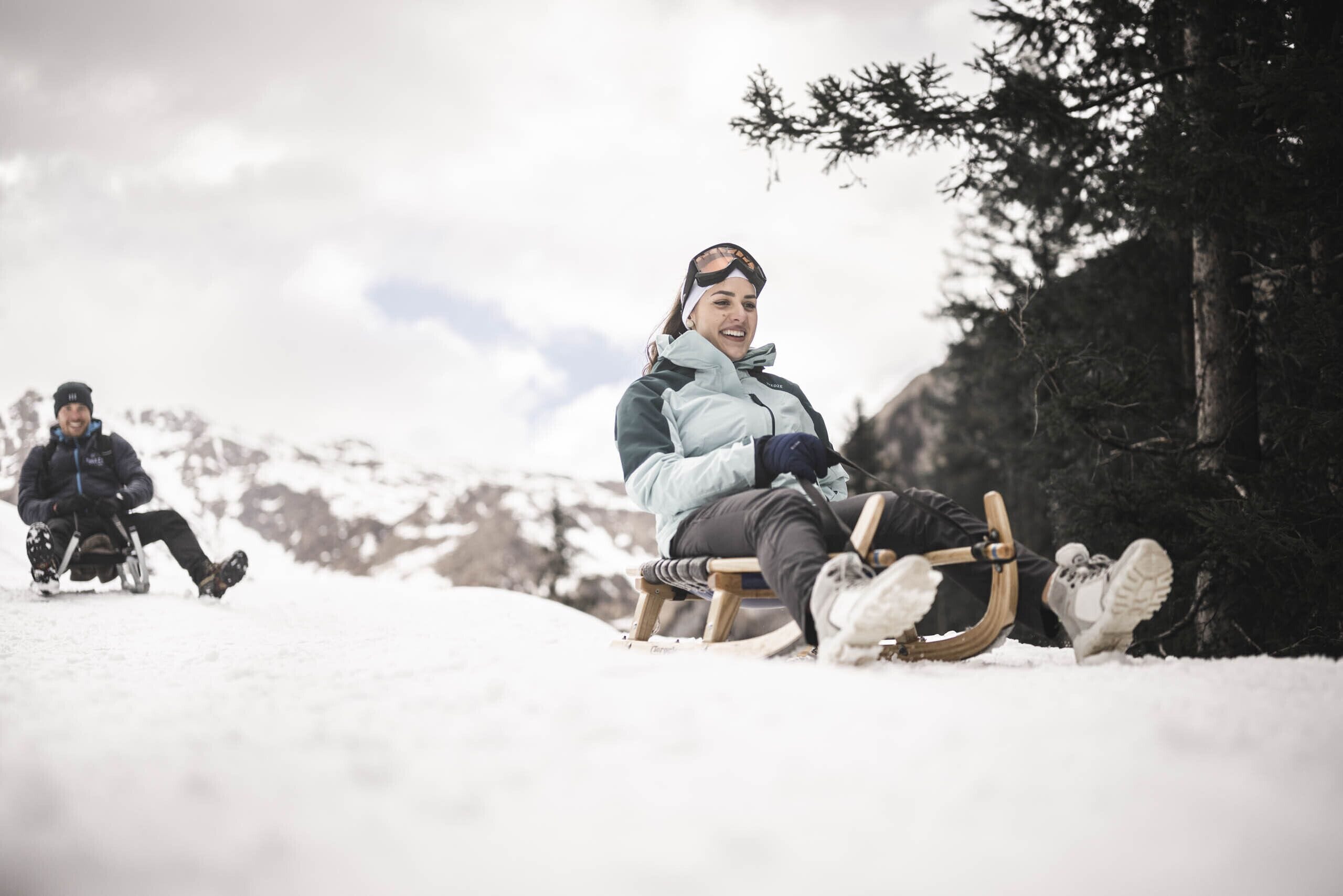 Una persona con una chaqueta azul claro y pantalones oscuros sentada en un trineo de madera en una pendiente nevada, con otra persona al fondo.