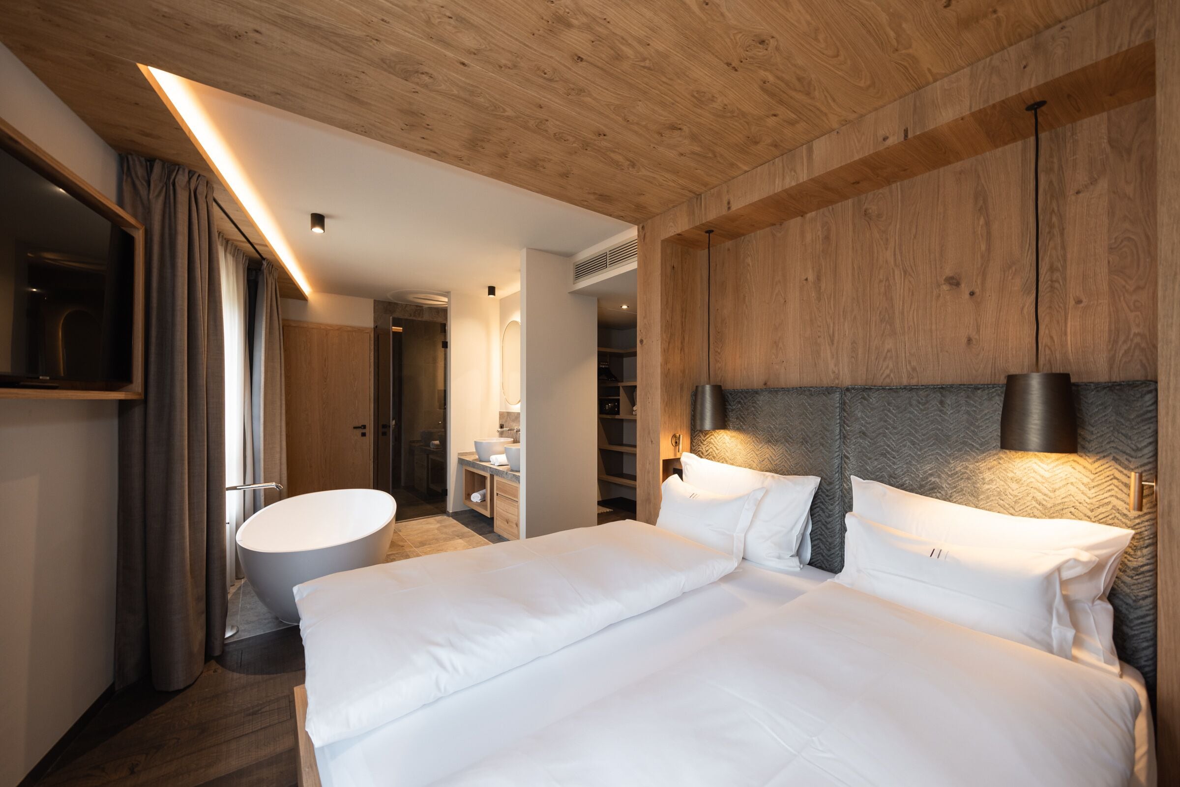 A modern hotel room with a large bed, a built-in bathtub, wooden ceiling, and a TV mounted on the wall.