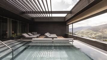 A modern indoor pool area with a skylight, lounge chairs, and a view of the mountains.