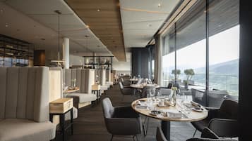 Un restaurant modern amb taules altes i seients, grans finestres que ofereixen una vista de la ciutat i les muntanyes.