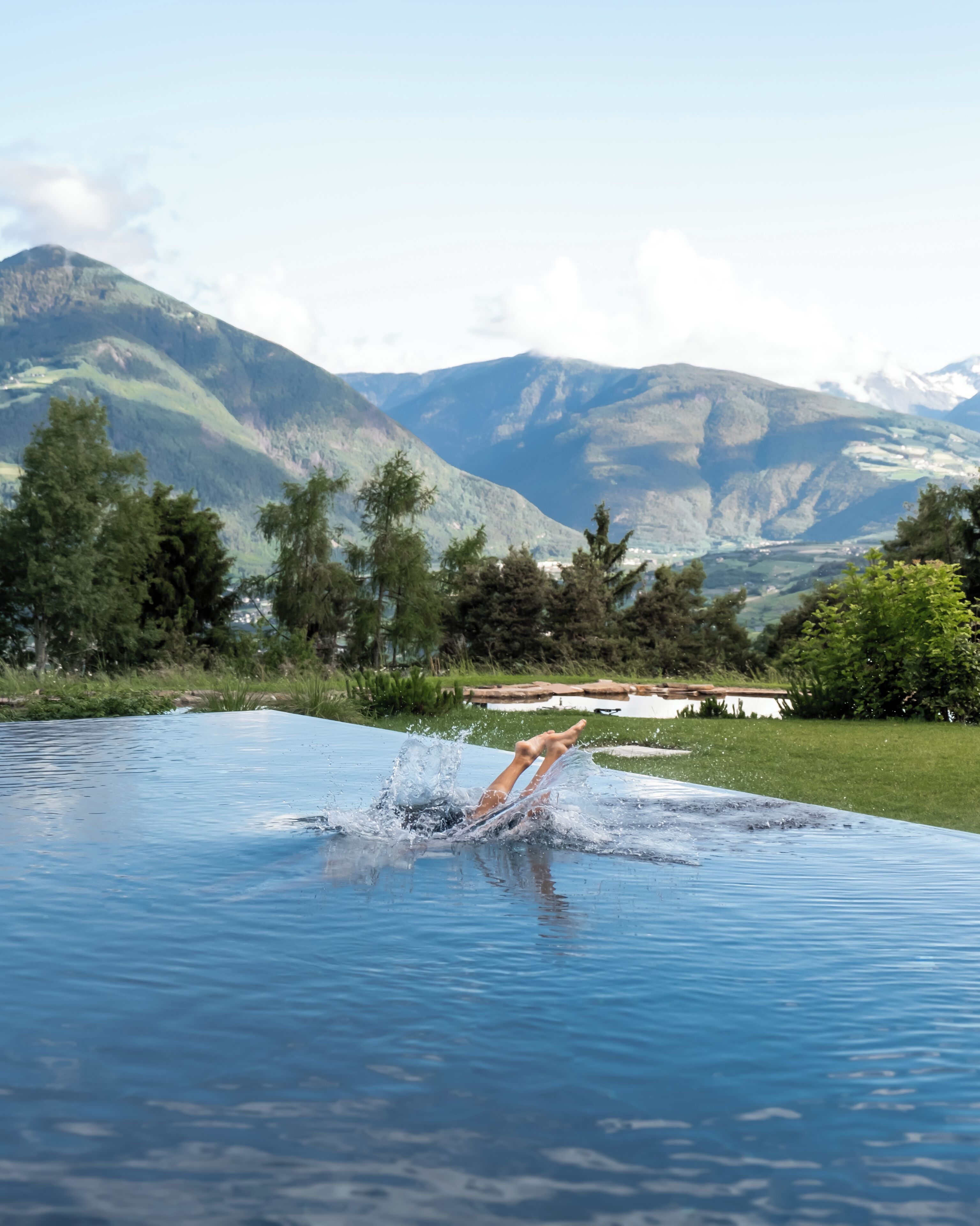A swimming pool with a person diving into the water, surrounded by mountains and greenery.