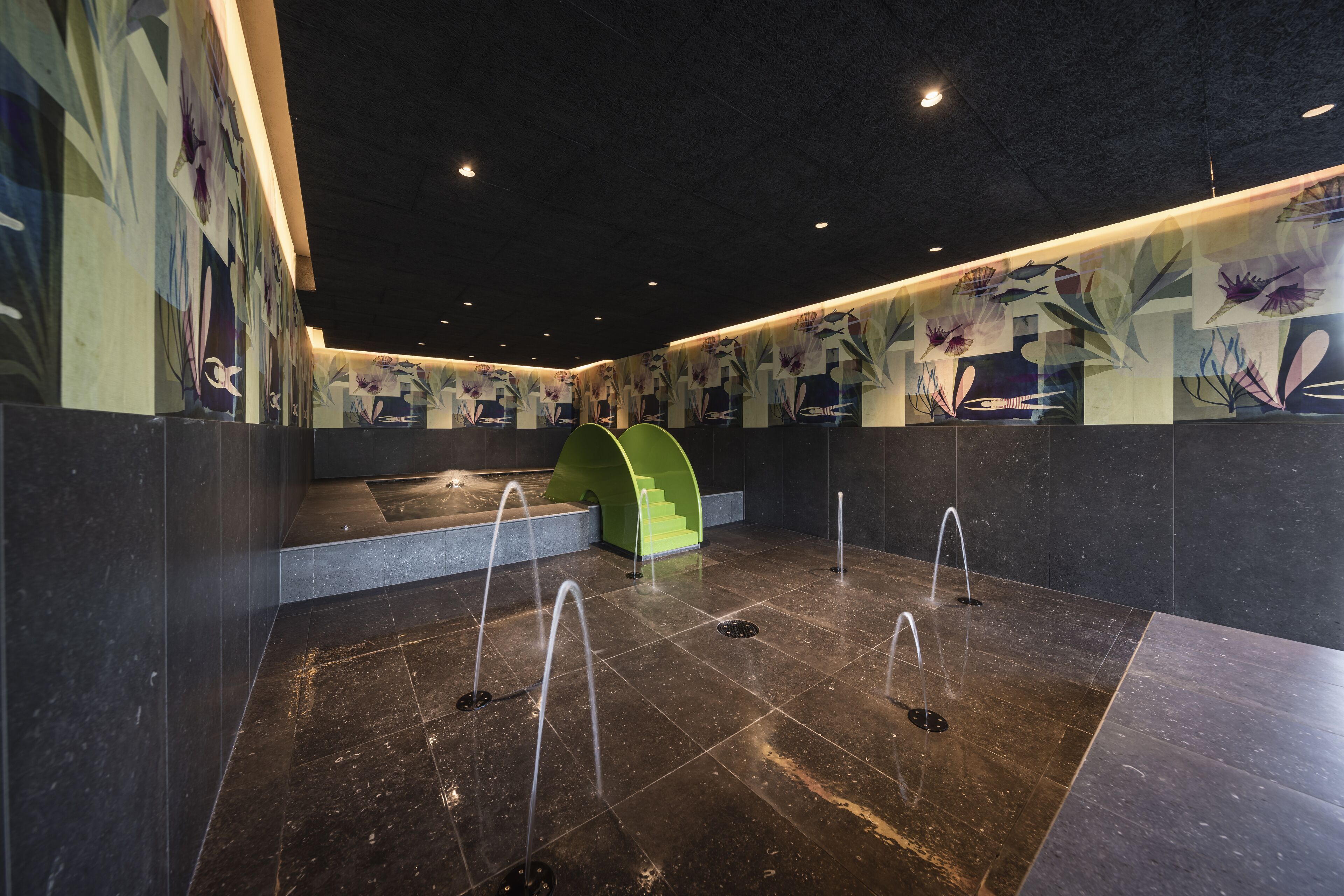 A modern indoor space with a dark tiled floor, green seating area, and a wall with abstract floral designs.