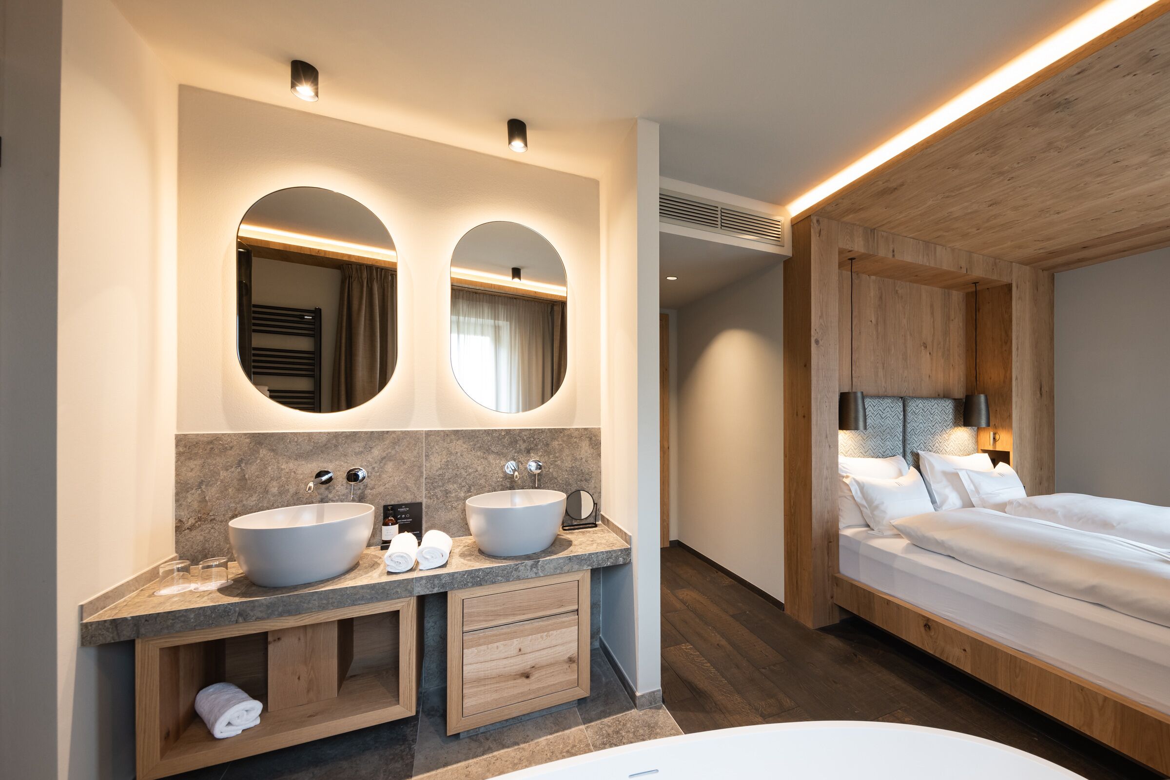 Une salle de bain moderne dotée d’un double lavabo, d’un meuble-lavabo, et d’une chambre séparée avec un lit et une table de chevet.