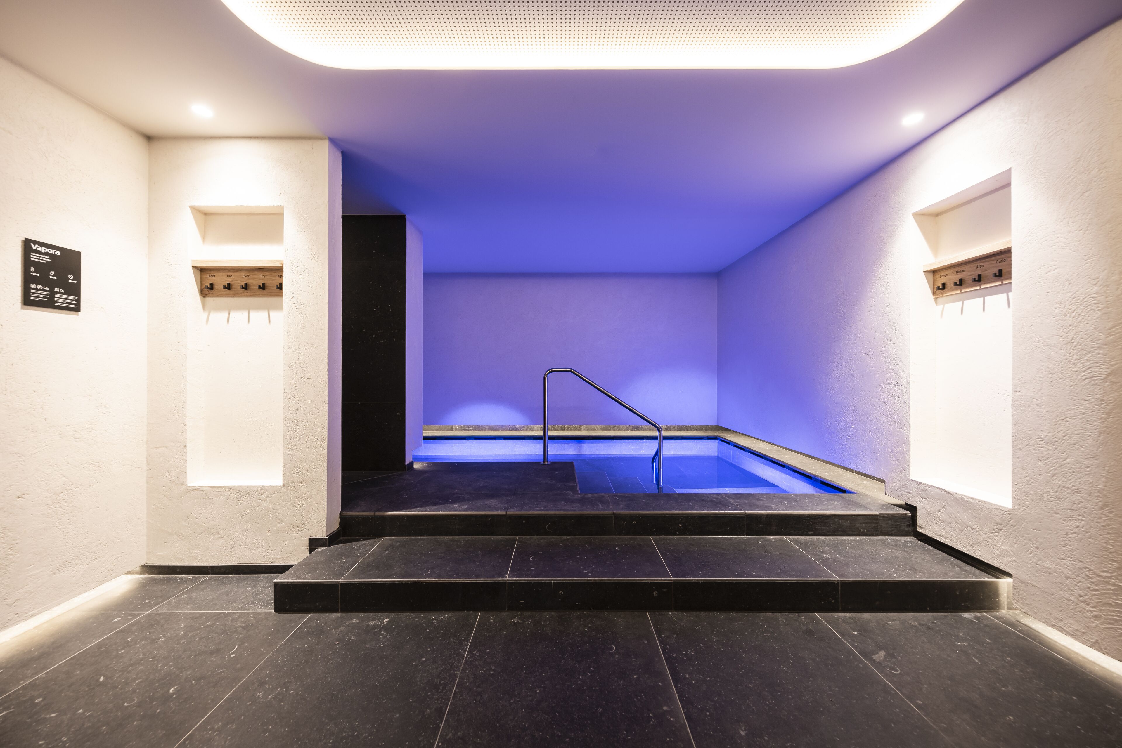 A modern indoor pool area with a blue-lit ceiling, a staircase leading to the pool, and a sign indicating a spa service.