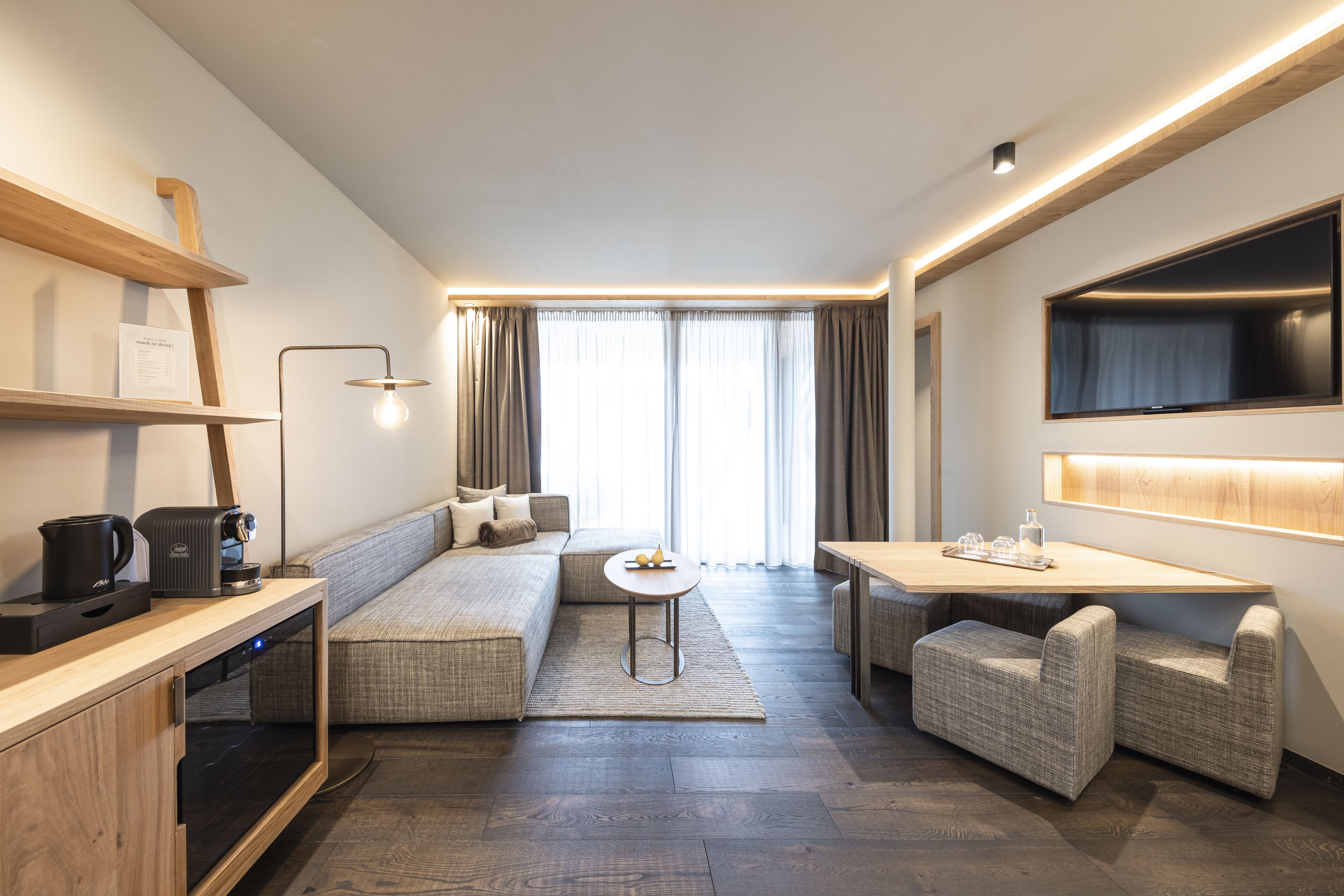 A modern hotel room with a sofa, a small table, a TV mounted on the wall, and a coffee machine on the counter.