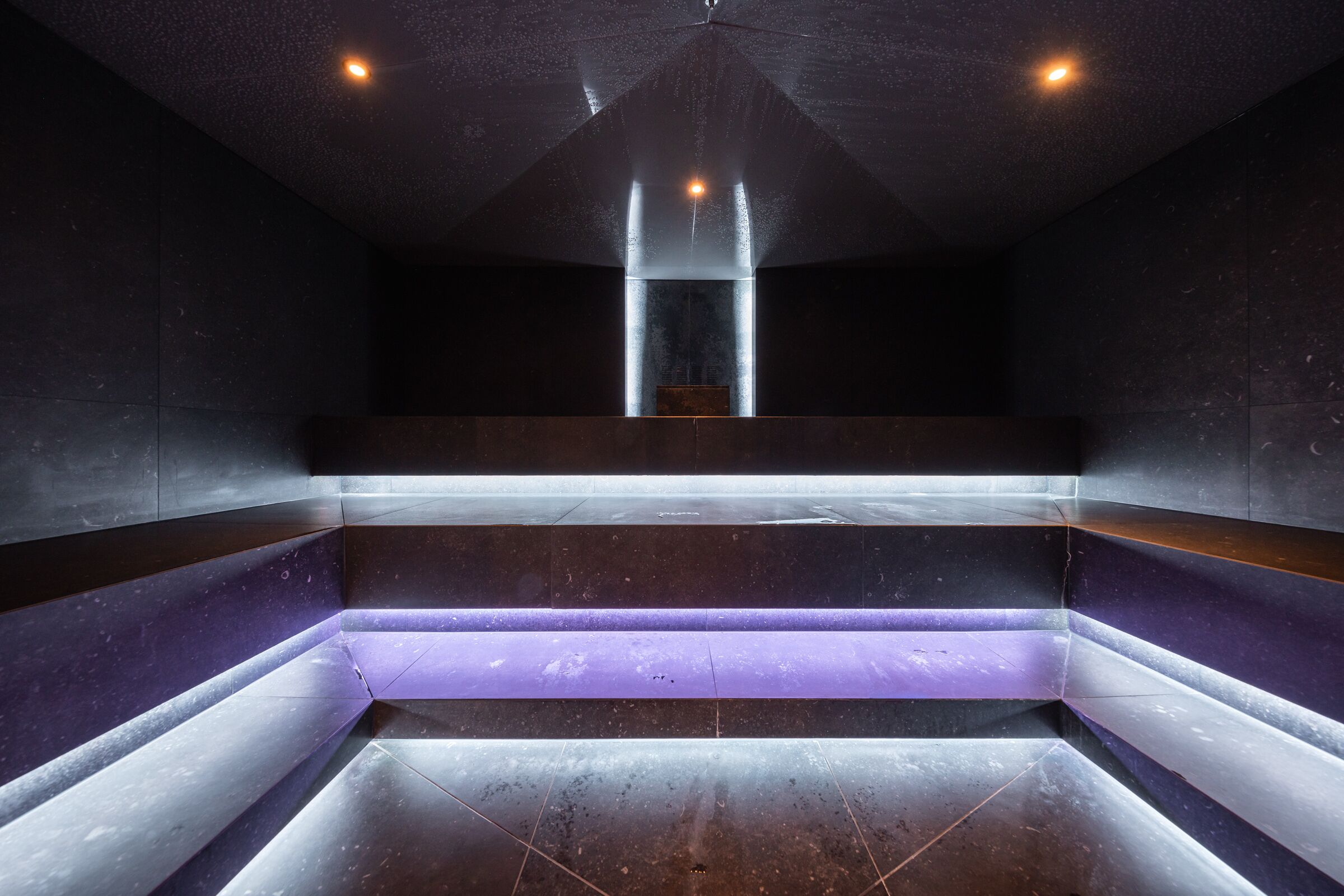 A modern, dimly lit interior with a central reflective water feature and a seating area with a purple-lit ledge.