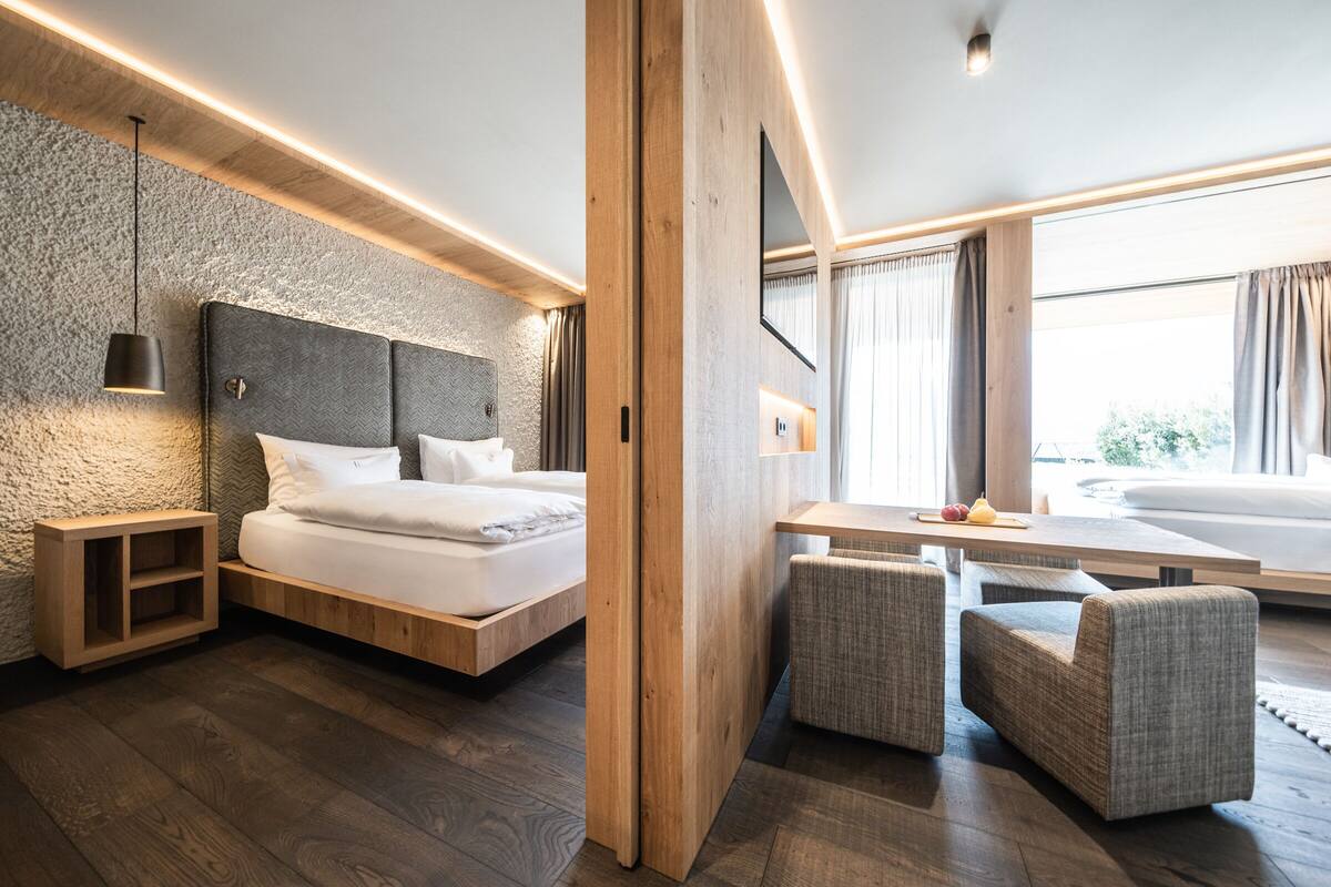 A modern hotel room with a large bed, a wooden headboard, a desk with two chairs, and a view of the outdoors.