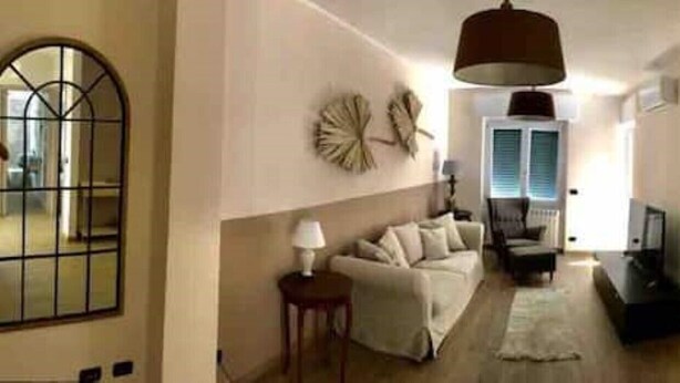 Een ruime woonkamer met een bank, een salontafel en een lamp. Er is een groot raam met jaloezieën, een deur en een trap die naar een andere kamer leidt.