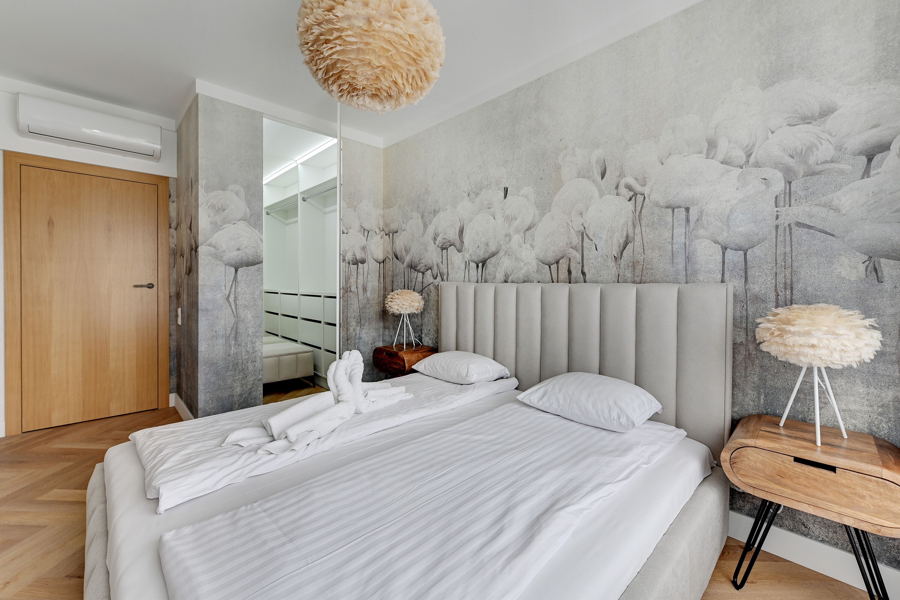 Une chambre moderne avec un grand lit, une tête de lit grise, des oreillers blancs moelleux et une fresque murale représentant des flamants roses.