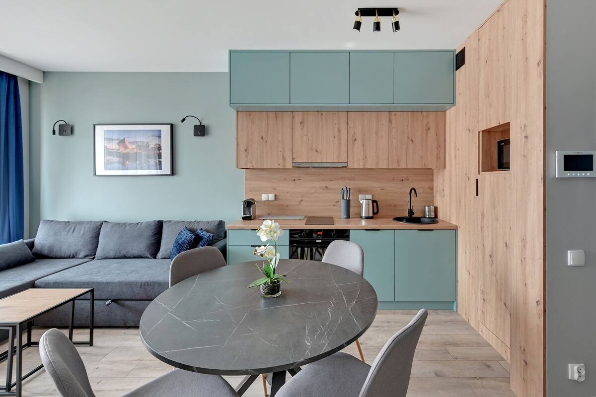 A modern living room with a grey sofa, a round dining table with chairs, and a kitchen area with wooden cabinets and a blue countertop.