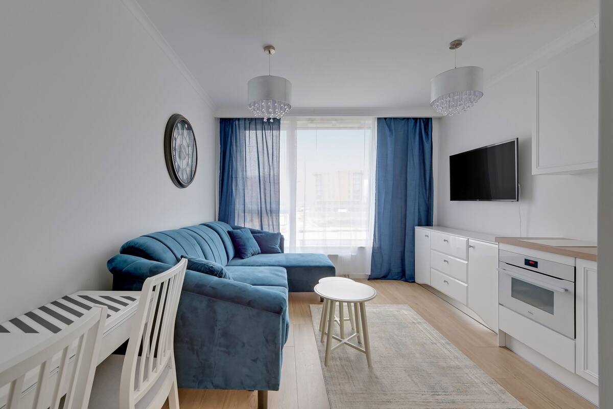 A modern living room with a blue sofa, a white dining table, and a kitchen area with a TV and cabinets.