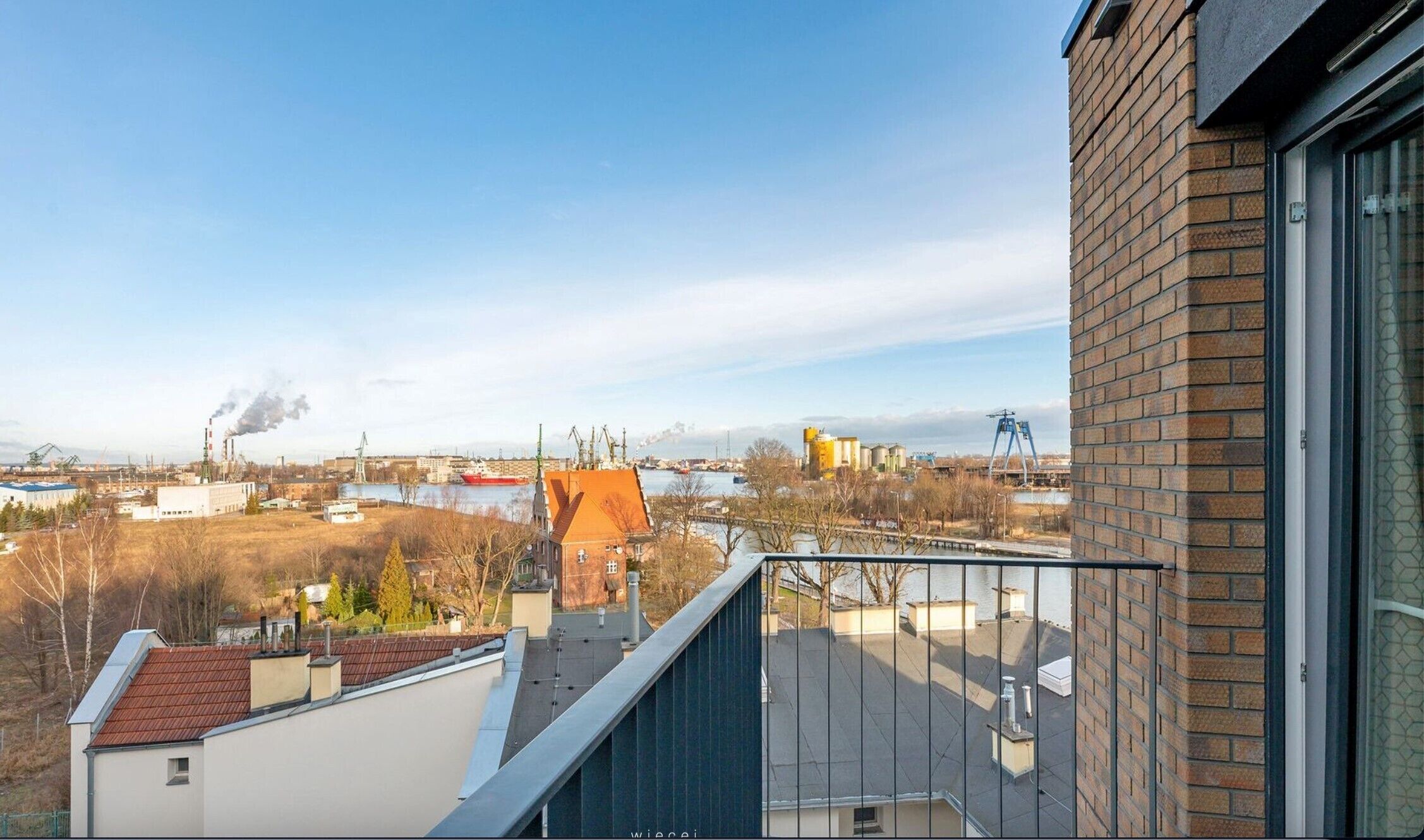 A residential building with a brick facade and a balcony overlooking a river with industrial structures in the background.