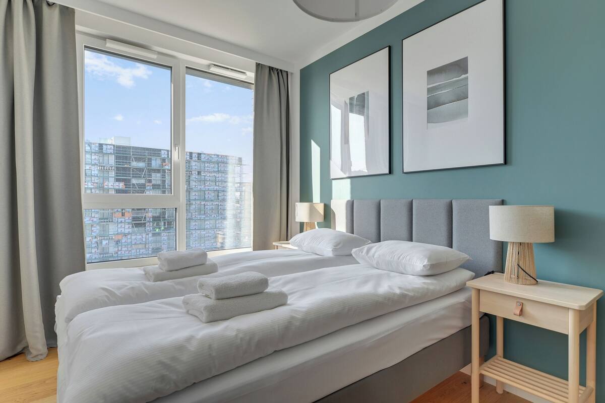A modern hotel room with a large bed, two bedside tables, a view of the city, and artwork on the walls.