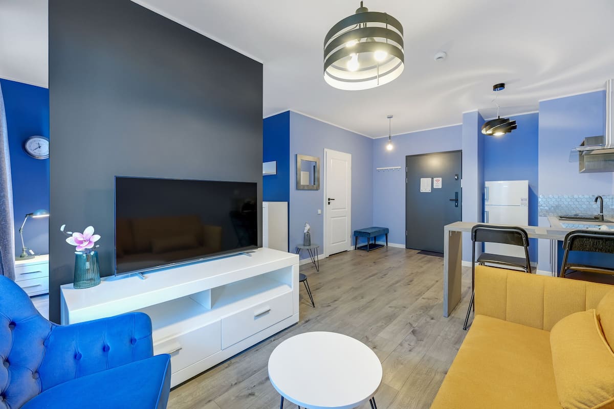 A modern living room with a flat-screen TV, blue and yellow sofas, a white coffee table, and a dining area with a small table and chairs.