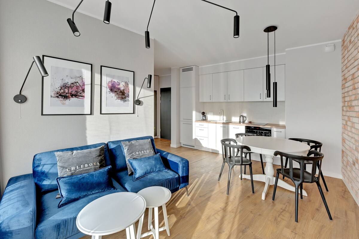 A modern living room with a blue sofa, white round tables, and black chairs. There is a kitchen in the background with white cabinets and a brick wall.