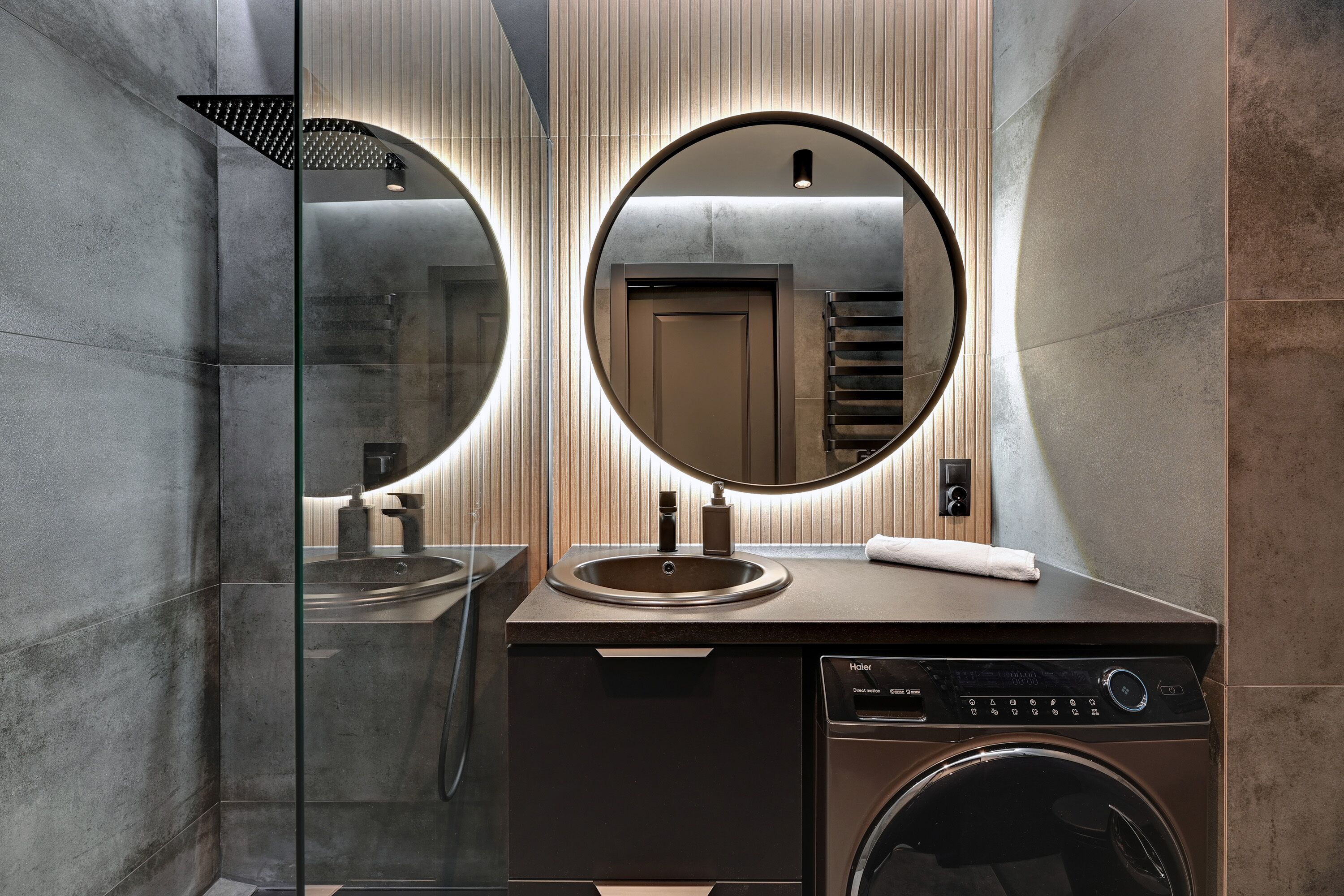 A modern bathroom with a glass shower enclosure, a round mirror, a sink, and a built-in washing machine.