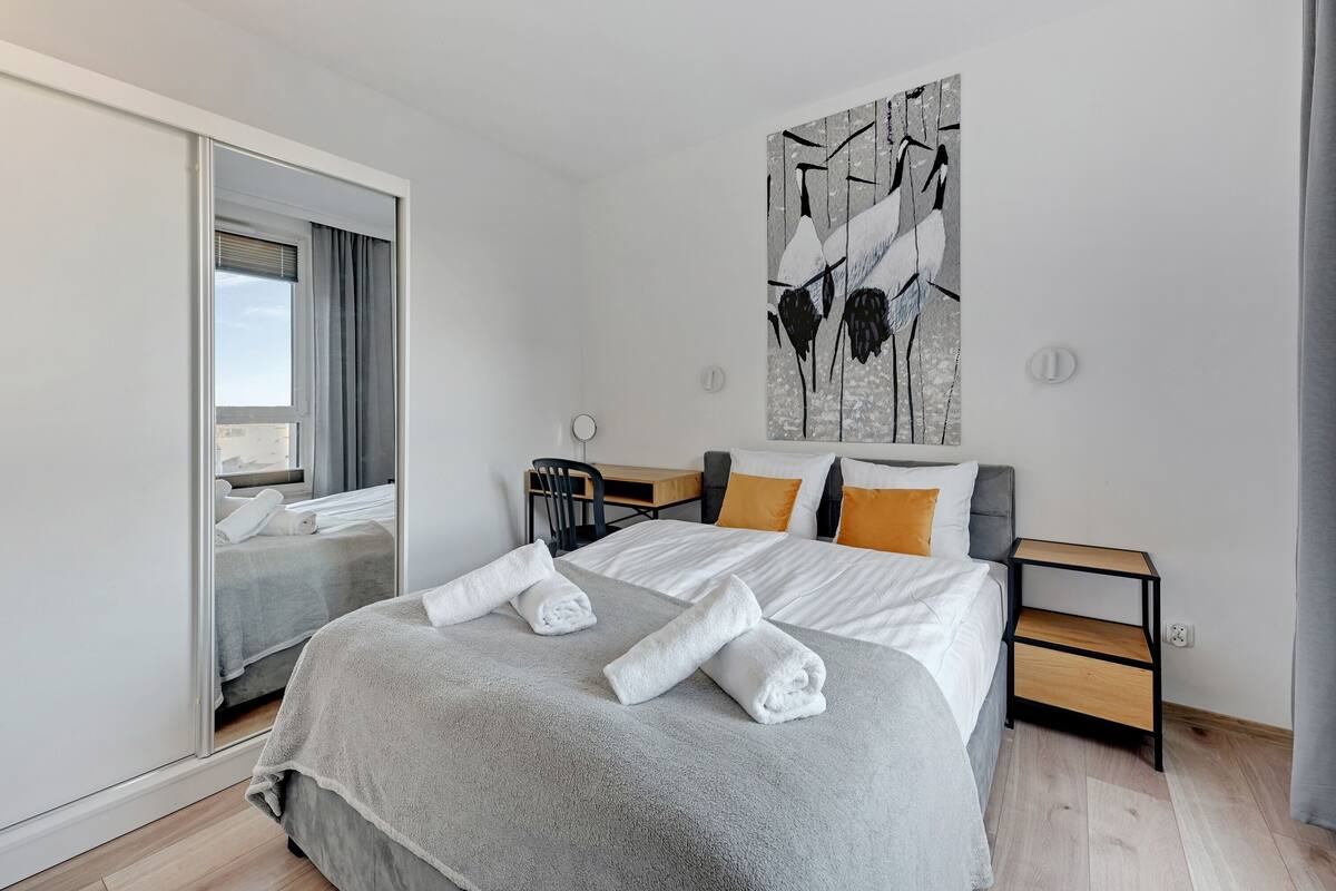 A modern hotel room with a large bed, a bedside table, a nightstand, a mirror, and a painting on the wall.