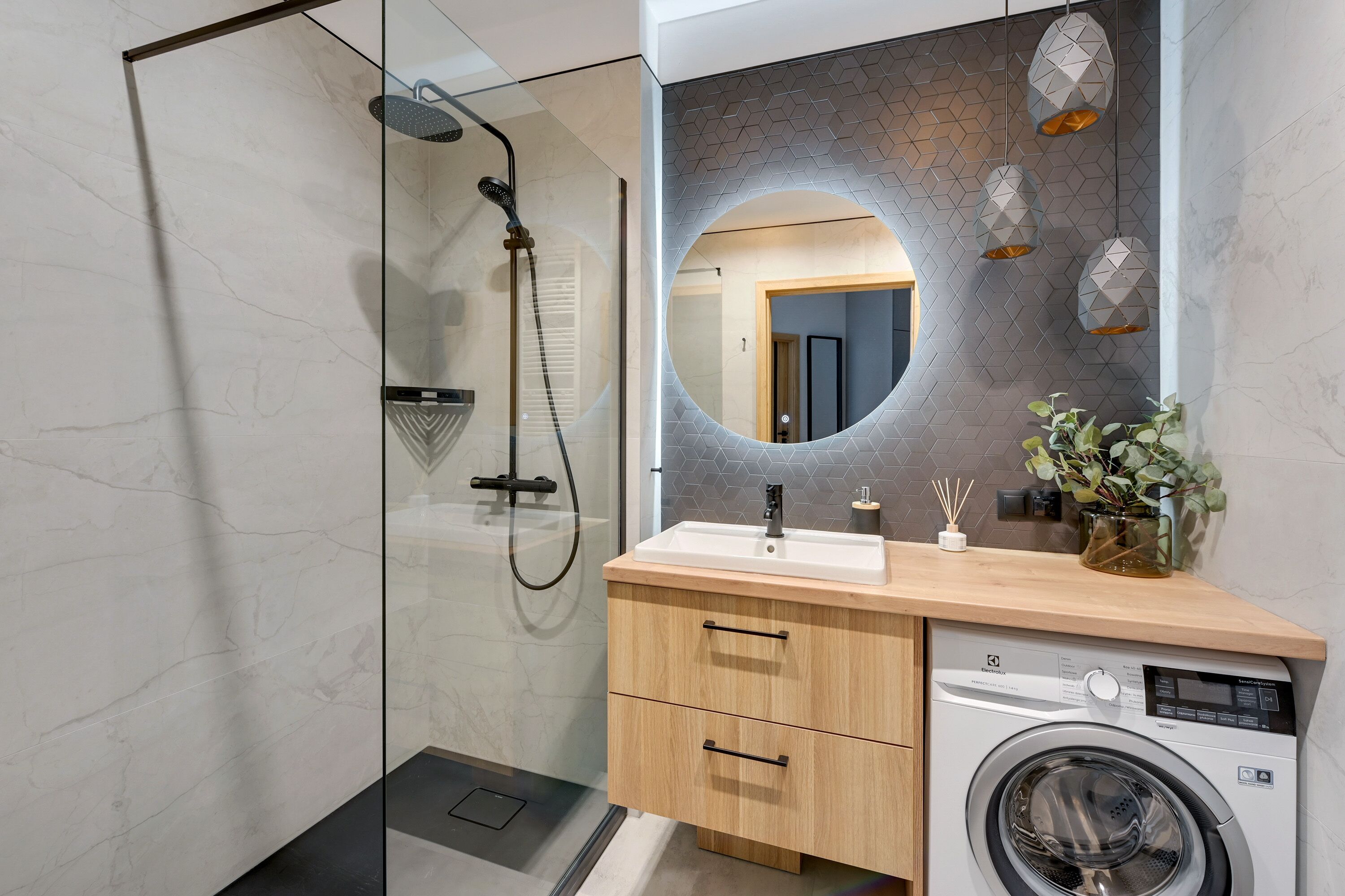 A modern bathroom with a glass-enclosed shower, a round mirror, a wooden vanity with a washing machine, and a geometric-patterned backsplash.