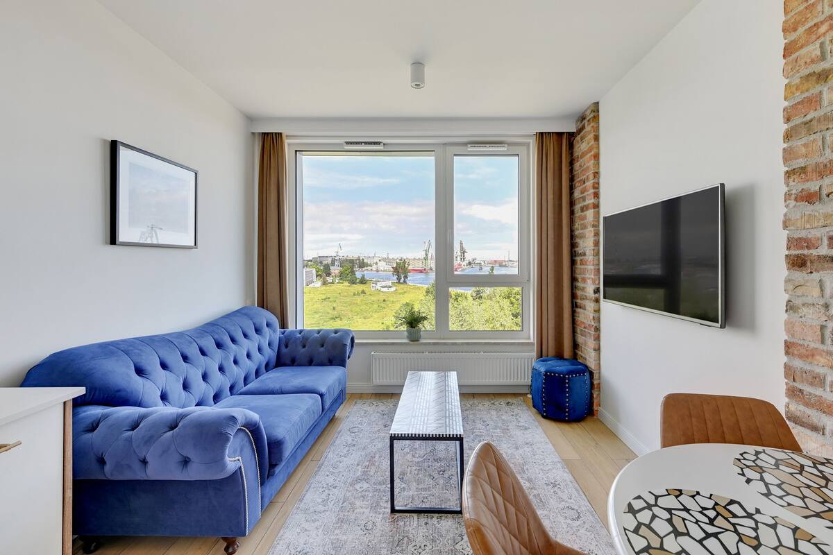 A modern living room with a blue sofa, a coffee table, a TV, and a view of a cityscape through large windows.